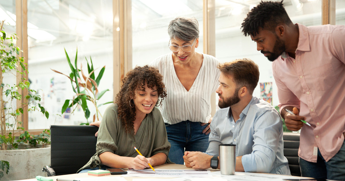 A group of diverse coworkers are gathered at a conference table in an office. The attention is on one coworker who is showing and explaining something to the others while using a pencil to point to a paper on the table.