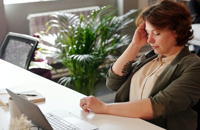 An image of a brunette woman wearing a green jack leaning back in her chair away from the laptop in front of her. She is rubbing her forehead with her eyes closed.