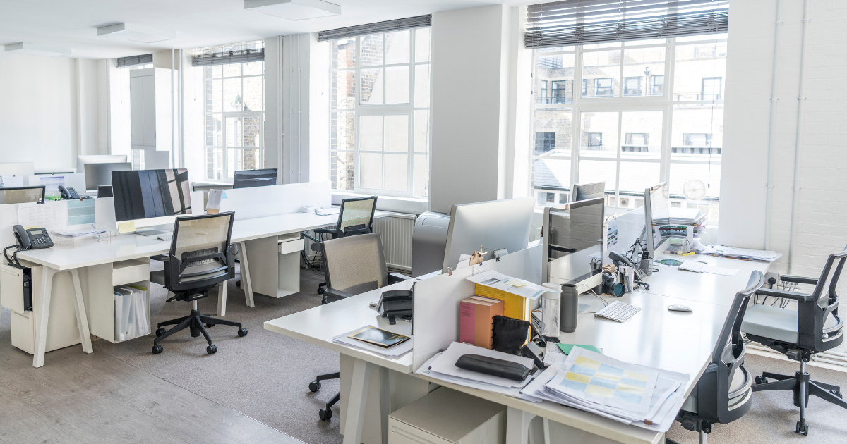 A bright Office with three large windows. There are two large tables with four workspaces on each table. Each workspace has a chair and a large computer monitor with a keyboard.