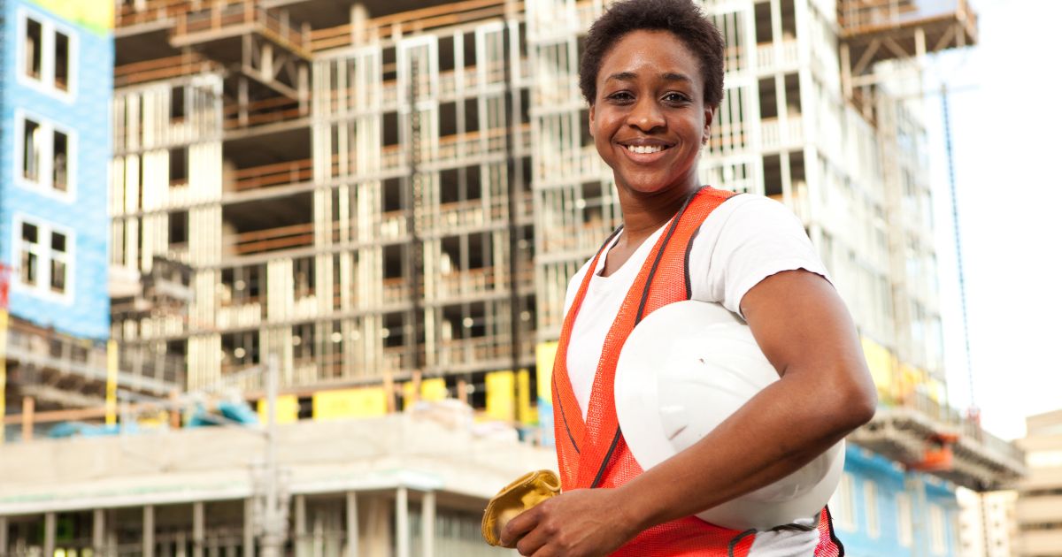 A construction worker, smiling, holding a hard hat under their arm. In the background is an unfinished building.