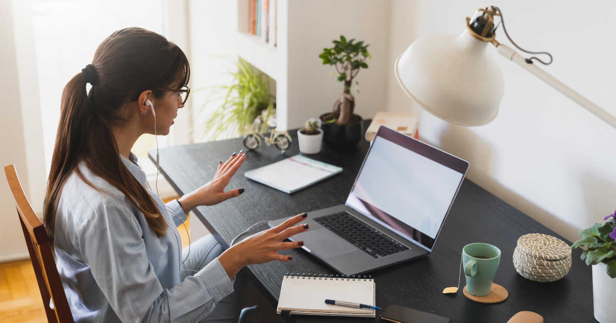 A person sitting at a black desk in a wooden chair with her hands in the air. There are plants, a coffee mug, a notepad, and a computer on the desk. Next to the desk is a bookshelf and a window with light shining in.