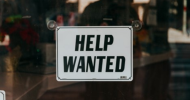 A black and white sign hanging in the window of a business that reads “Help Wanted.” 
