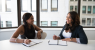 Two people in an office smiling at each other while conducting an interview and taking notes. One is writing notes in a white notebook and the other is smiling and responding. 