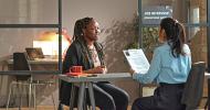 Two people sitting across a desk from each other in an office during a job interview. The interviewer is holding the applicant’s resume.